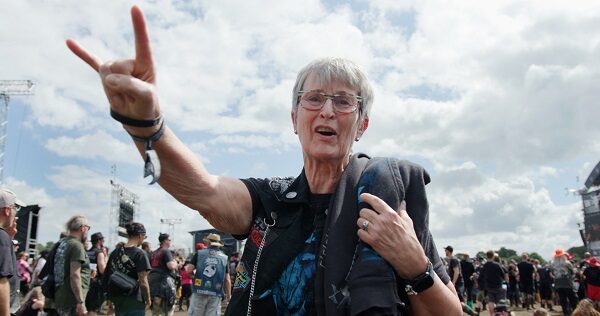 Eine ältere Frau steht vor einer Menschenmenge und zeigt die sog. "gehörnte Hand" in die Luft